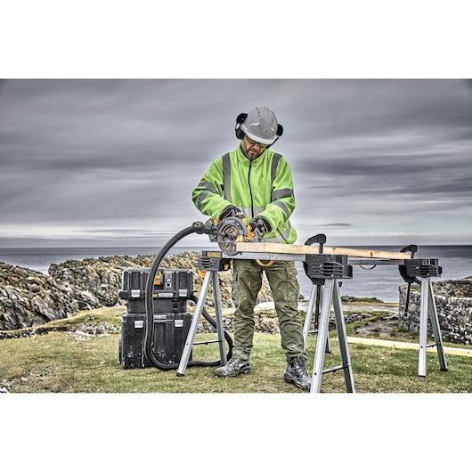 A person wearing a high-visibility jacket, helmet, and ear protection is using a DEWALT circular saw to cut wood outdoors. The wood is placed on two sawhorses, and there are DEWALT tool boxes nearby. The scene is set against a rocky coastal landscape under a cloudy sky.