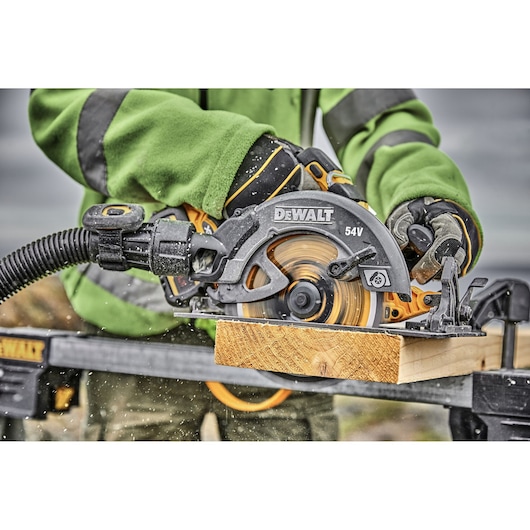 A person wearing gloves and a green jacket uses a DEWALT 54V cordless circular saw to cut a piece of wood, with sawdust and debris visible as the blade spins.