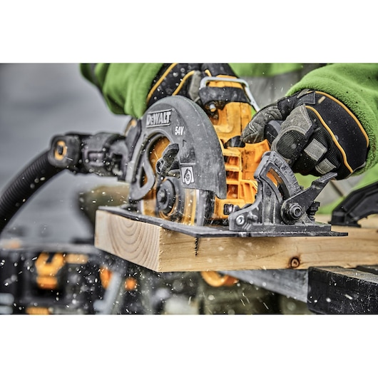 A close-up image of a DEWALT 54V cordless circular saw cutting through a wooden plank, operated by a person wearing green and protective gloves. Sawdust is flying from the cut.