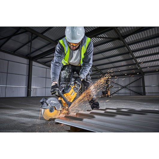 Worker using DEWALT DCS690N saw to cut metal roofing, sparks flying, in an industrial warehouse.