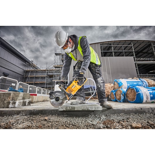 Person using a DEWALT power saw to cut concrete at a construction site, wearing safety gear and a high-visibility vest.