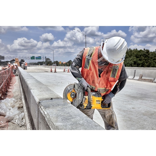 Worker uses DEWALT saw to cut concrete barrier on highway construction site, wearing safety gear and helmet.