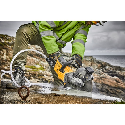 A person wearing a green jacket and protective gloves uses a DEWALT DCS690X2 power tool to cut through a wet concrete surface outdoors, with rocky terrain and water in the background.