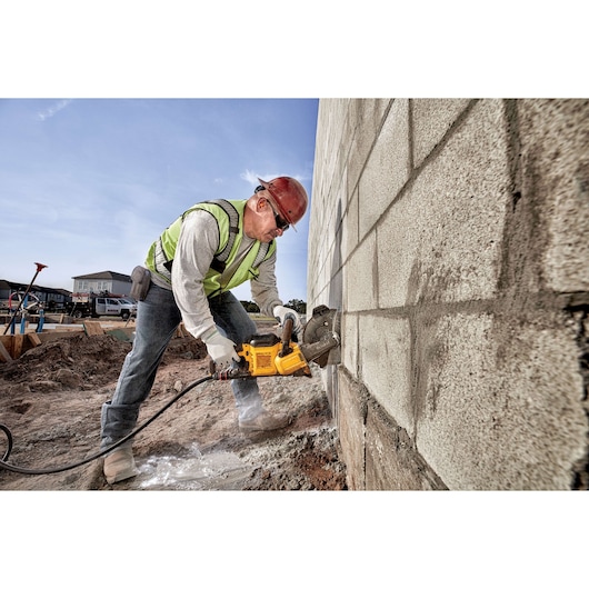 A construction worker wearing a safety vest and helmet is using a yellow DEWALT power tool to cut into a concrete wall at an outdoor construction site.