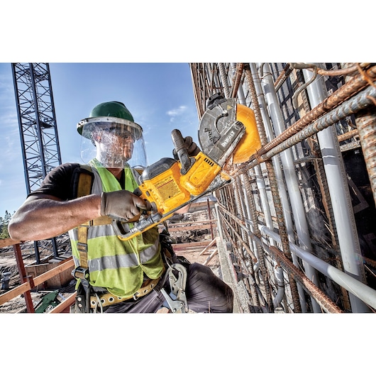 A construction worker wearing safety gear uses a yellow DEWALT cordless cutting tool to cut through metal rebar at a building site. The worker is equipped with gloves, a reflective vest, helmet, and face shield.
