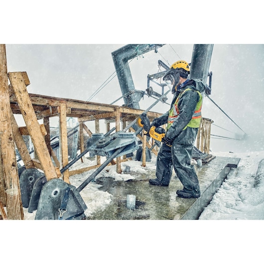 A construction worker wearing weatherproof clothing, a safety vest, and a helmet operates a DEWALT power tool outdoors in snowy conditions. The worker is cutting through a thick metal rod attached to a wooden structure, with visible snow and industrial equipment in the background.