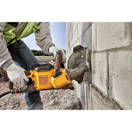 A person using a DEWALT DCS690X2 power tool to cut through a concrete block wall. The person is wearing gloves, jeans, and a safety vest, and the tool is generating visible dust during operation.