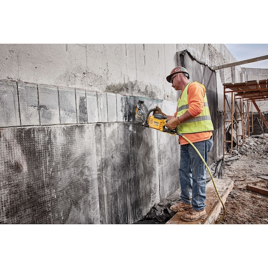 A construction worker using a DEWALT power cutter (SKU: DCS690X2) to cut into a concrete wall at an outdoor construction site. The worker is wearing a safety vest, gloves, jeans, boots, and a hard hat.