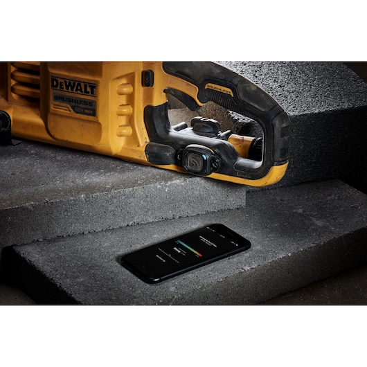 A close-up of a yellow DEWALT 60V MAX brushless power tool resting on concrete blocks, with a smartphone displaying a control or monitoring app placed nearby.