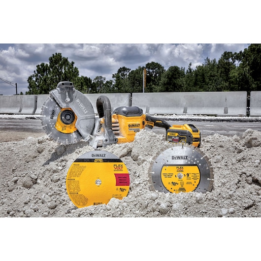 A DEWALT cordless concrete cutting saw with FLEXVOLT battery is placed on a pile of light-colored construction material outdoors. Two DEWALT cutting discs, one for metal and one for concrete, are displayed in front of the saw. The scene is at a construction site with barriers and trees in the background.