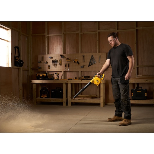 Person uses a DEWALT DCV100 blower to clear dust in a workshop with tools and wooden workbench.