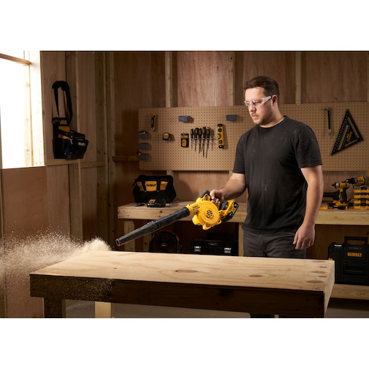 Person using a DEWALT cordless blower to clean sawdust from a wooden workbench in a workshop.