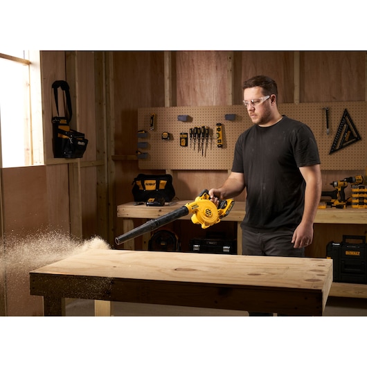 Person using a DEWALT DCV100 blower to clean sawdust off a wooden workbench in a workshop.