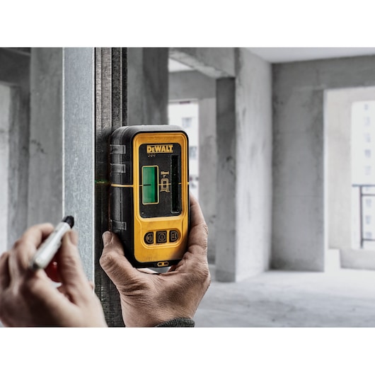 A person is using a DEWALT DE0892G green laser detector to measure a level line on a metal beam in an unfinished indoor construction site. The hand is holding the device and marking the beam with a pen.