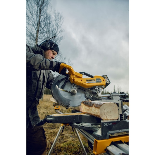 A person operates a DEWALT DHS780N miter saw outdoors, cutting a piece of wood on a portable workbench. The weather appears overcast and there are trees in the background.