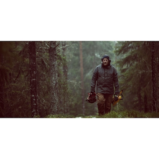 A person wearing protective clothing and headphones is walking through a forest while carrying a DEWALT chainsaw and a safety helmet.