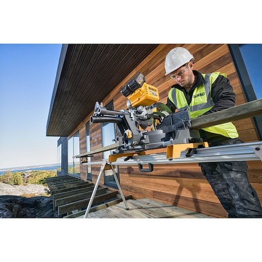 A construction worker using a DEWALT sliding miter saw (model DHS780N) with a yellow and black design to cut a wooden board outdoors beside a wooden building. The worker is wearing a hard hat and a high-visibility safety vest.