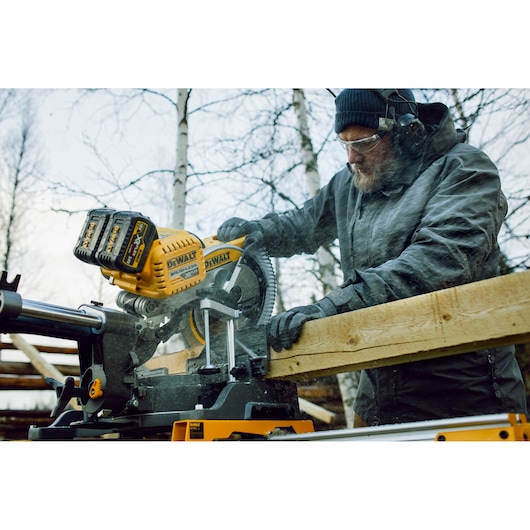A person using a DEWALT DHS780N cordless mitre saw to cut a wooden beam outdoors. The person is wearing protective gloves, a jacket, and ear protection while operating the saw. The DEWALT yellow saw is prominently visible, and there are trees in the background.