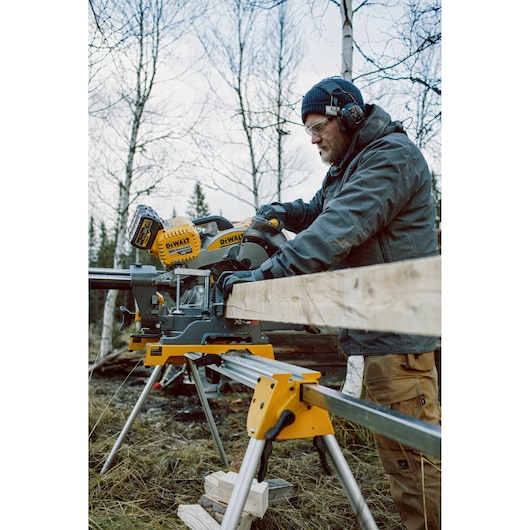 A person wearing protective gear operates a DEWALT DHS780N cordless mitre saw outdoors, cutting a wooden plank supported by a stand. The scene is set in a wooded area with leafless trees.