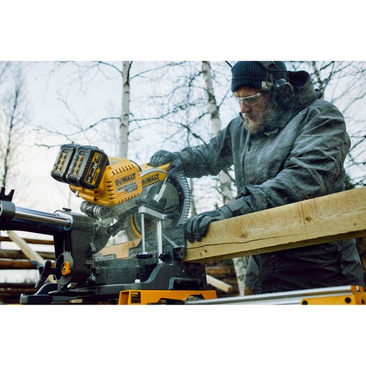 A person wearing protective clothing is using a DEWALT DHS780T2 cordless sliding miter saw to cut a wooden board outdoors. The saw is yellow and black with visible dual battery packs, and wood dust is being generated during the cut.