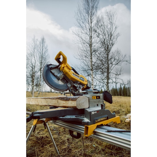 DEWALT miter saw model DHS780T2 set up outdoors on a workbench, cutting a wooden beam. Leafless trees and a cloudy sky are visible in the background.