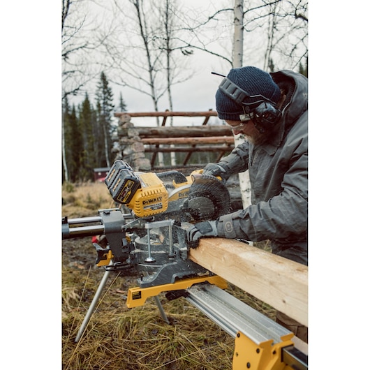 A person using a DEWALT DHS780T2 miter saw to cut a wooden beam outdoors, wearing safety earmuffs, gloves, and a beanie. The saw is set up on a stand in a woodland area.