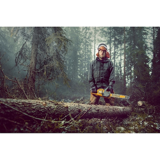 A person wearing protective gear stands in a forest holding a DEWALT chainsaw, next to a fallen tree trunk. The forest is misty and dense with tall trees.