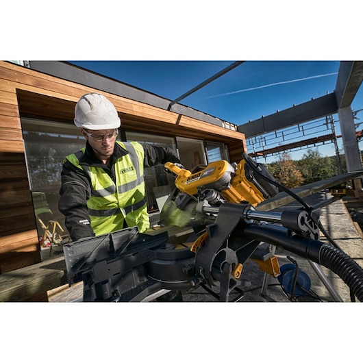 A construction worker wearing a safety vest and helmet operates a DEWALT DHS780T2 miter saw outdoors on a building site. The saw is cutting a wooden plank, and the work area includes a partially constructed timber building under a clear blue sky.