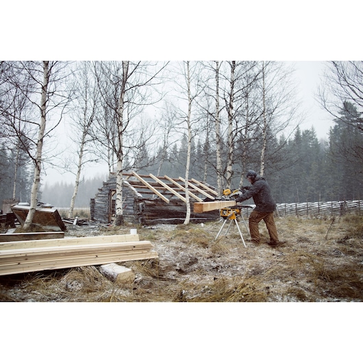 A person using a DEWALT miter saw outdoors in a snowy, wooded area, cutting a large piece of wood. There is a partially constructed wooden structure and stacks of lumber nearby.