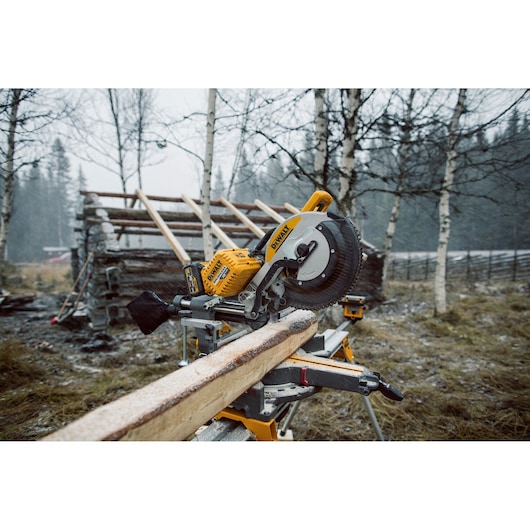 A DEWALT DHS780T2 miter saw is set up outdoors on a stand, cutting a frost-covered wooden board. The scene is in a snowy, wooded area with bare trees and a partially built wooden structure in the background.