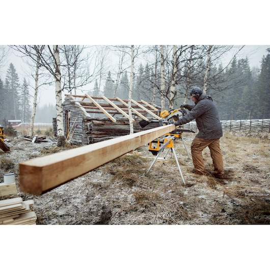 A person using a DEWALT sliding compound miter saw outdoors to cut a long wooden beam, with a partially built wooden structure and trees in the background.