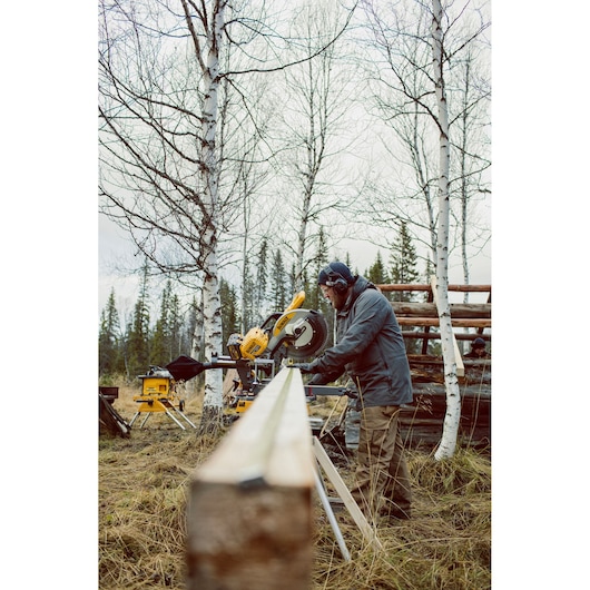 A person using a yellow DEWALT miter saw (SKU: DHS780T2) outdoors in a forested area with birch trees. The saw is set up on a stand, and the person is cutting a long wooden plank while wearing ear protection and outdoor clothing.