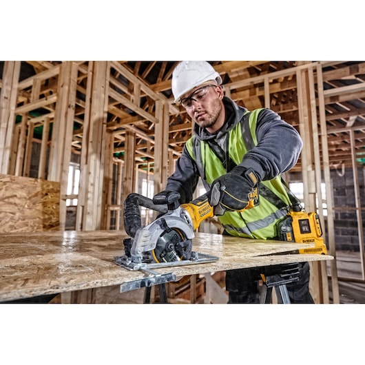 Worker uses a DEWALT power saw to cut wood in a building under construction, wearing safety gear.