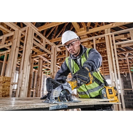 Worker uses DEWALT circular saw on wood at construction site, wearing safety gear and reflective vest.