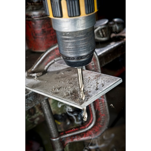 Close-up of a DEWALT drill bit drilling through a metal sheet clamped in place, metal shavings around the hole.