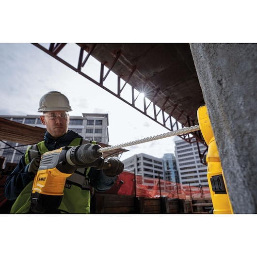A person wearing a safety helmet and vest uses a DEWALT rotary hammer drill with a long drill bit on a construction site with buildings in the background. The person's face is blurred for privacy.