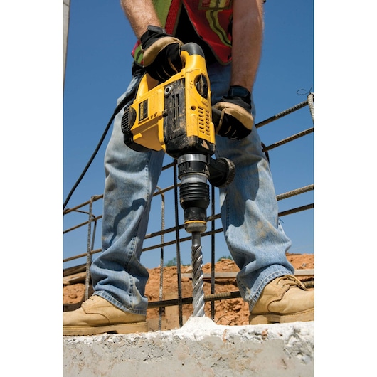 Close-up of a person using a yellow DEWALT power drill to drill into concrete at a construction site. The person is wearing work gloves, jeans, safety boots, and a reflective vest.
