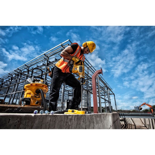 Worker uses DEWALT drill on concrete at construction site, wearing safety gear and helmet.