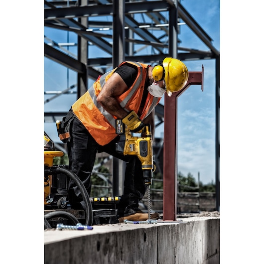 A construction worker wearing a yellow hard hat, orange safety vest, ear protection, and a face mask uses a cordless DEWALT rotary hammer drill on a concrete surface at an outdoor construction site, with steel framework visible in the background.