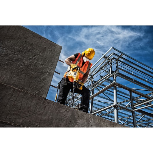 A construction worker wearing a yellow hard hat, orange safety vest, and face mask is using a yellow DEWALT power tool to drill into concrete at a building site with metal framework structures under a blue sky.