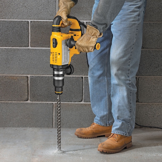 A person wearing gloves and work boots is using a yellow DEWALT rotary hammer drill to drill into a concrete floor. The drill has a long masonry bit and the worker is standing next to a gray concrete block wall.