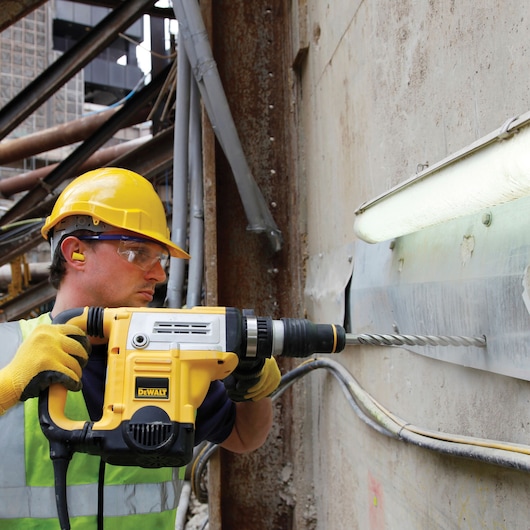 A construction worker wearing a yellow hard hat, safety gloves, and high-visibility vest operates a DEWALT rotary hammer drill to bore into a concrete wall at a construction site.