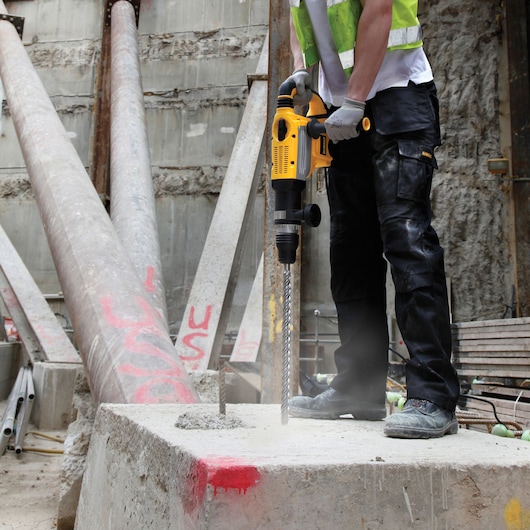 A person wearing protective clothing uses a yellow DEWALT rotary hammer drill to drill into a concrete block at a construction site.