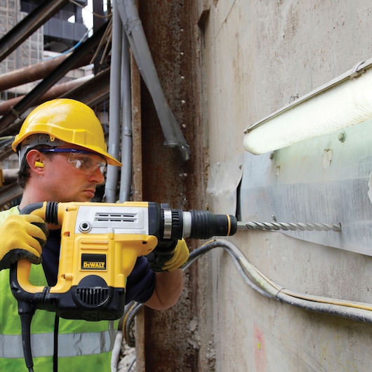A construction worker wearing a yellow hard hat and gloves is using a DEWALT rotary hammer drill to drill into a concrete wall on a construction site. The worker is equipped with ear protection and a safety vest.