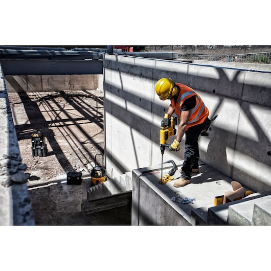 A construction worker wearing a yellow safety helmet and orange reflective vest is using a DEWALT power drill to drill into a concrete wall on a construction site. Various tools and equipment are visible around the site.