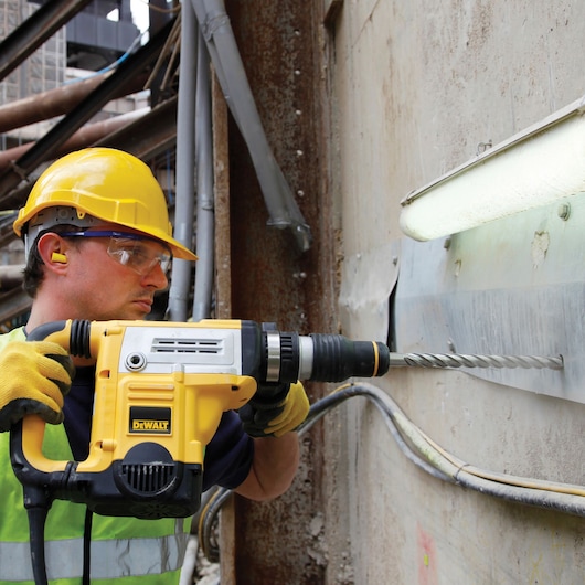 A construction worker wearing a yellow helmet, ear protection, and gloves is using a DEWALT rotary hammer drill to drill into a concrete wall at a construction site.