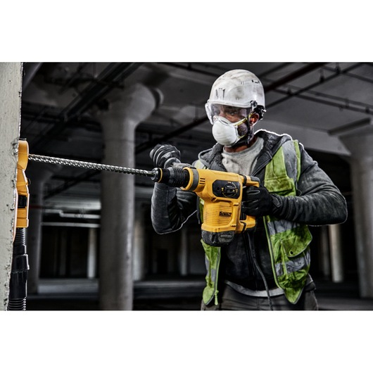 A construction worker wearing safety gear and a reflective vest is using a yellow DEWALT hammer drill with a long drill bit to drill into a concrete wall in an industrial setting.