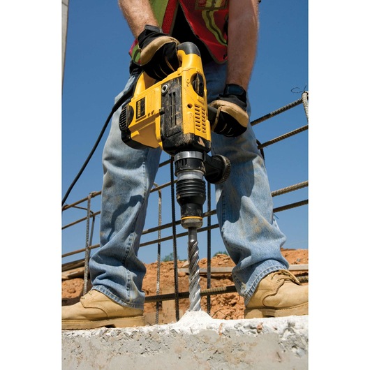 A person using a DEWALT yellow power drill with a long bit to drill into concrete at a construction site. The person is wearing gloves, jeans, boots, and a safety vest.