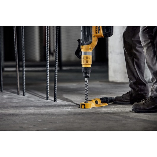 A person is using a DEWALT rotary hammer drill with a drill bit to drill into a concrete floor. Metal rebar rods are visible in the background, and the work appears to be taking place at a construction site.
