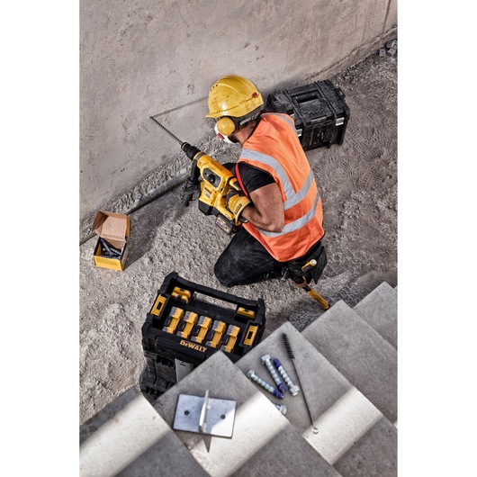 A construction worker wearing a yellow hard hat, orange safety vest, and ear protection is using a DEWALT power drill to drill into a concrete wall. Nearby, there are DEWALT toolboxes, a box of drill bits, and construction materials on the ground. Some stair steps with bolts and a brush are visible in the foreground.